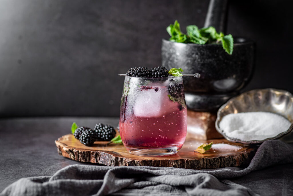 a scene whit a dark background with a balckberry mojito in the front over a butchers block. In the back some of the ingredients of the mojito